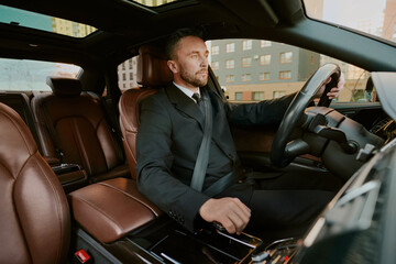 Caucasian middle aged man driving luxury car, wearing formal suit, holding steering wheel with left hand, looking focused through windshield, sitting in front seat