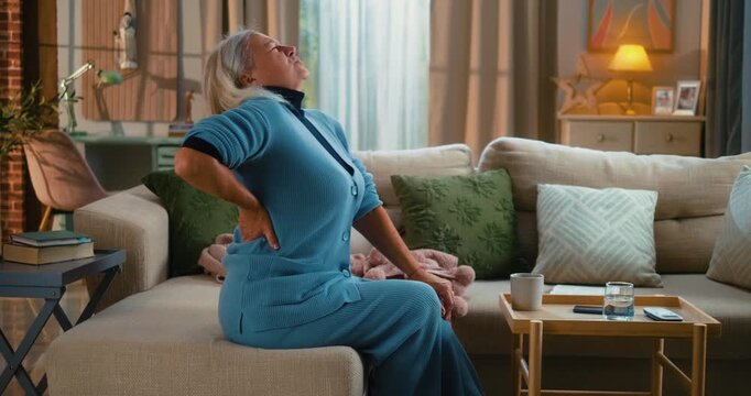 Middle-aged woman suffering from severe lower back pain, grimacing and rubbing her back while struggling to stand up from a sofa. Concept of discomfort, tension, and physical strain.