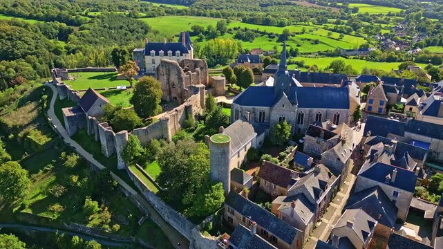 Aerial pull-out drone shot showing the medieval town of Sainte-Suzanne in Mayenne, France, with its stone castle, church, ramparts, and rooftops surrounded by lush green countryside and rolling hills.