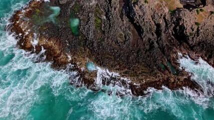 Fixed drone shot showing ocean waves hitting the rocky shore of Smaills Beach, with seabirds flying over the turquoise water. - Powered by Adobe