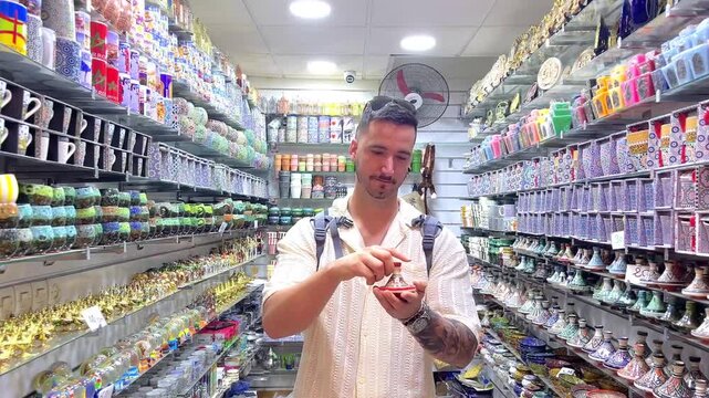 White european young male tourist with a backpack choosing a traditional moroccan tagine in a souvenir store during his vacation in Marrakech's souk looking traditional handicrafts and gifts in Africa
