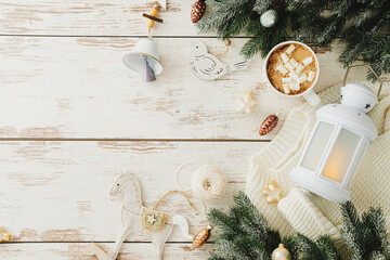 Elegant Christmas flat lay with glowing white lantern, warm knitted sweater, evergreen branches, bell, wooden bird ornament, and hot chocolate on a white wooden surface.