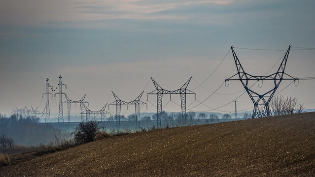 Row of high-voltage power line towers across rolling fields. Rural landscape with repeating steel structures.