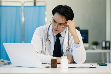 Stressed Asian male doctor sitting at a desk with a laptop, feeling tired and anxious while working in a hospital office.