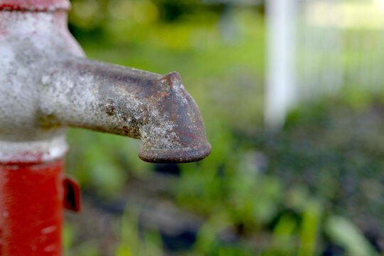 Close-up of the rusty, weathered spout of an old, red and white hand pump against a blurred, green outdoor background.