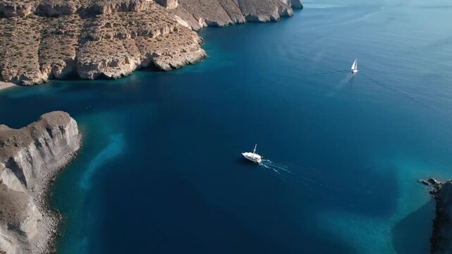 An breathtaking aerial drone shot captures two pristine vessels navigating the tranquil, deep blue waters of a secluded bay, flanked by rugged, towering cliffs and a small, inviting cove. A white sail
