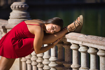 Young woman, brunette, gymnast, Latina, wearing a red dress, stretching with her face resting on her leg on a stone railing. Concept: youth, sports, gymnastics, stretching, contortionism.