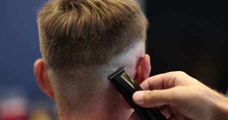 Barber hand with electric clipper provides haircut to man client occiput in salon. Master uses modern hairdo tool serving man in barbershop