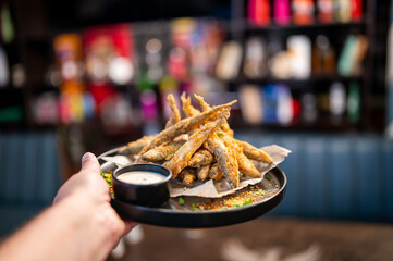 Hand holding a black plate of golden-brown, deep-fried small fish (likely anchovies or smelts) served with a white dipping sauce, on a parchment-lined plate with a blurred restaurant background.