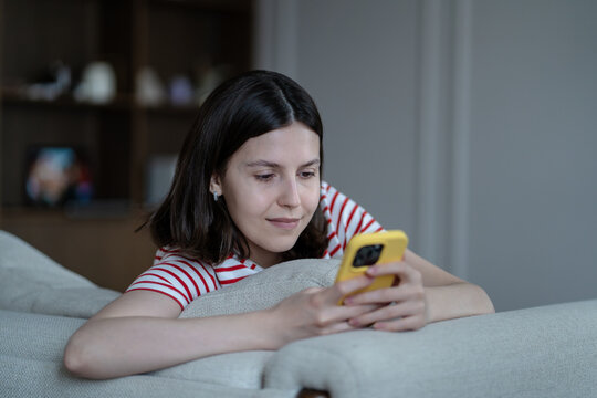 Contented smiling teen girl rest on couch absorbed in smartphone screen, scrolling social media or chatting online. Serene happy adolescent browsing webs, texting friends, immersed in internet world. - Powered by Adobe