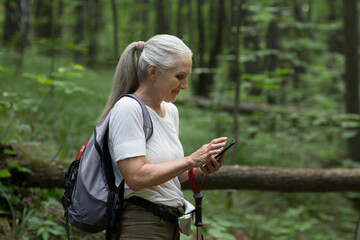 senior woman  with  a backpack uses a mobile phone standing on in the summer forest. copy space. mental health. Slow life.  
