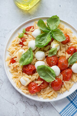 Orzo pasta with mini mozzarella, roasted cherry tomatoes and green basil, vertical shot on a white stone background, middle closeup