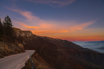 Mountain road curving along dramatic ridges at sunset, with colorful sky and misty valleys in the distance, capturing peaceful travel, nature beauty and highland landscapes.