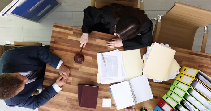 Female judge taps gavel sitting opposite businessman at wooden table. Entrepreneur breaks corporate laws and gets punishment by attorney upper view