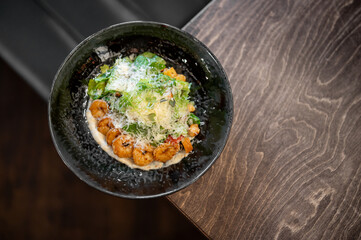 A close-up, top-down view of a gourmet shrimp salad in a dark bowl, featuring fresh green lettuce, grated parmesan cheese, and a creamy sauce on a rustic wooden table
