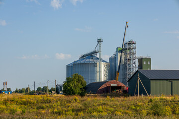 Large silos and a granary are situated in an agricultural area with a crane preparing for storage operations on a sunny day