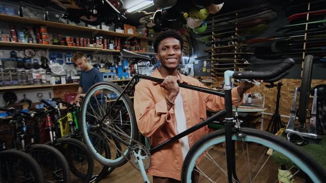 Young Black man working in repair shop, lifting bicycle to check its handlebar and steerer tube, smiling at camera, his Caucasian colleague working in background