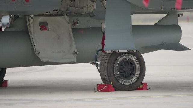 Side view of military aircraft main landing gear with wheel secured by chocks and visible external fuel tank