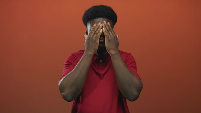 Man covers eyes with hands in studio orange wall wearing red polo shirt, hands over face visible gesture; shyness introspection.