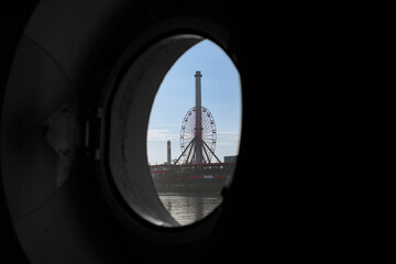 View of a Ferris wheel from a circular window on a boat during daytime at a waterfront amusement park