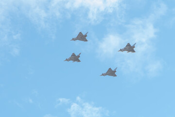 Four jet fighters are flying in a dense group in the blue sky under light clouds.