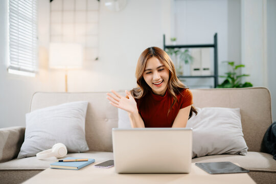 Cheerful Asian woman wearing headphones smiling and waving hands during online video call at home. Modern communication, friendship, and remote connection concept.