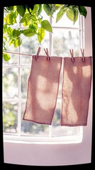 Two pieces of fabric are hung on a clothesline with clothespins in front of a window, with sunlight and green leaves visible.