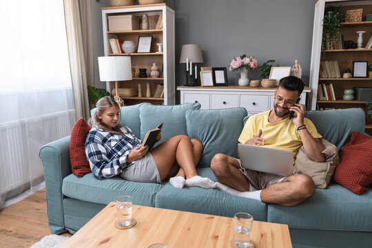 Relaxed couple winding down on couch, she enjoys a book while he works on a laptop, sharing smiles, easy conversation and balanced downtime that blends quiet reading with light digital leisure at home