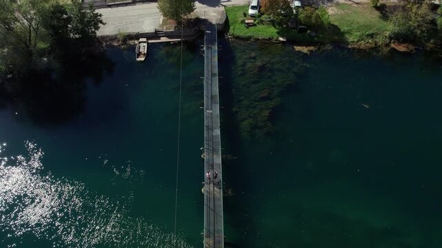 Drone footage of a wooden footbridge crossing the Una River near Bihac, Bosnia and Herzegovina. Clear turquoise water, reflections, and people walking above the river in calm nature.