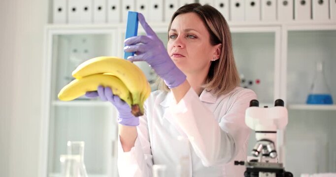 Woman laboratory assistant analyzes bananas using special scientific device. Blonde laboratory worker observes readings on nitrate tester monitor