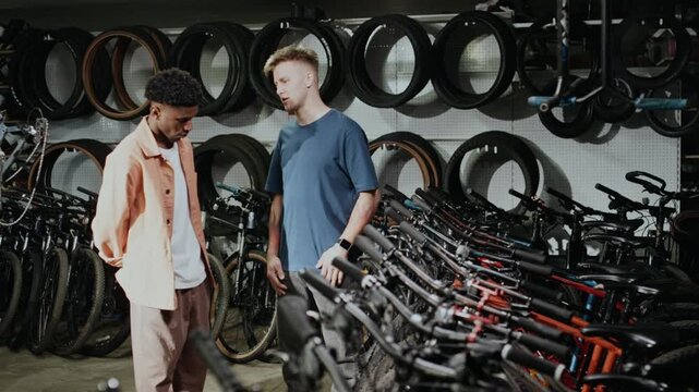 Young Caucasian shop owner describing technical specifications of bicycles, helping young Black customer to choose one