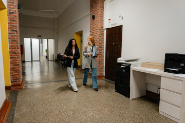 Two women walk and chat in a bright office corridor with brick pillars and a printer desk.