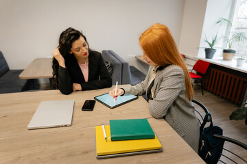 Two colleagues work together in a modern office, one writing on a tablet while the other observes.