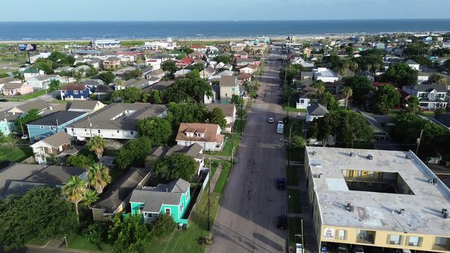 Classic Galveston residential neighborhood with colorful, elevated homes along deep green trees lines perpendicular street leading toward sandy shoreline of Stewart Beach area, Texas Gulf Coast
