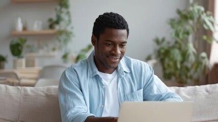 A cheerful young man with a warm smile is deeply engaged with his portable computer, comfortably seated on a cozy sofa in a bright, modern home interior. The scene radiates a sense of relaxed producti