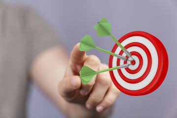 Close-up of a hand pointing at a dartboard with darts in the center, symbolizing success,...