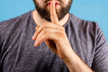 Close-up of a bearded man shushing with finger on lips, isolated on blue. Concept of silence,...