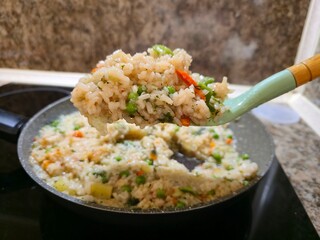 Close-up of a spoonful of steaming rice pilaf with vegetables being lifted from a nonstick frying pan on a kitchen stove