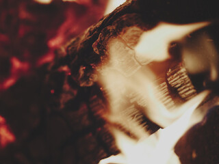 Close up of charred log burning in fireplace, flames passing over the wood, glowing red embers in blurred background, warm cozy atmosphere, dramatic focus on texture and fire details.