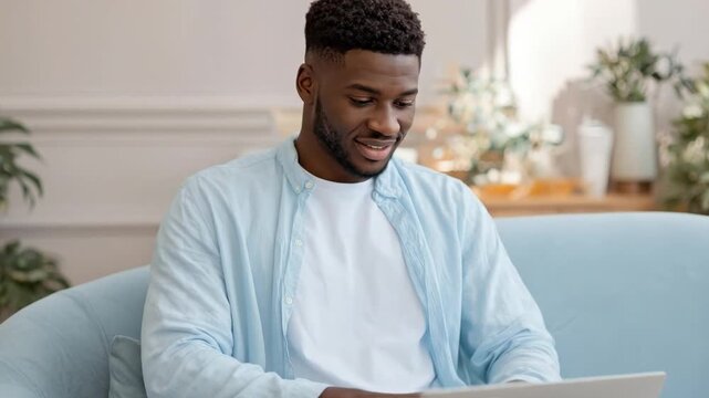 A smiling young person of African descent sits comfortably on a modern, light blue sofa, fully immersed in their activity on a laptop computer. Their cheerful expression and focused gaze downwards ind