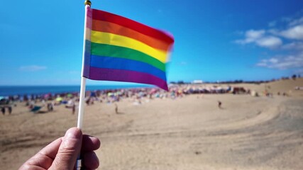 Person's hand holding a small waving LGBT rainbow pride flag in slow motion with a crowded beach in the background during the Winter Pride celebration in Maspalomas, Gran Canaria - Powered by Adobe