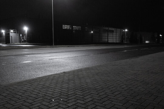 A quiet industrial area at night, illuminated by street lights, with wet asphalt and empty sidewalks. The scene creates a sense of isolation and silence.