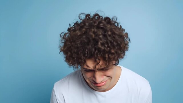 A young man with curly hair expresses profound sadness and emotional distress, captured against a clean, light blue studio background. His face is contorted with grief, eyes squeezed shut, and mouth o