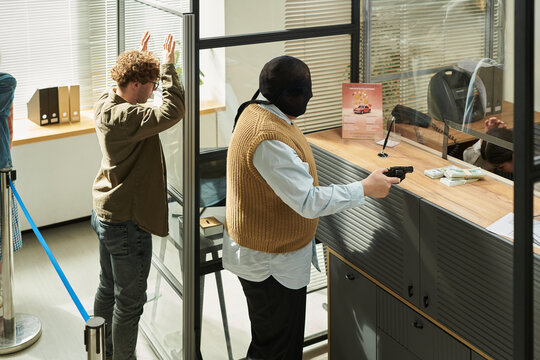 Caucasian young adult man wearing mask holding handgun and demanding money from bank teller while Caucasian young adult man standing with hands raised against glass partition