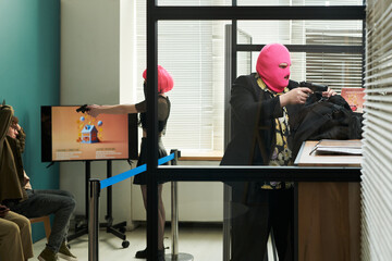 Two young adults wearing pink balaclavas robbing bank, one aiming handgun at seated people while other opening bag at counter