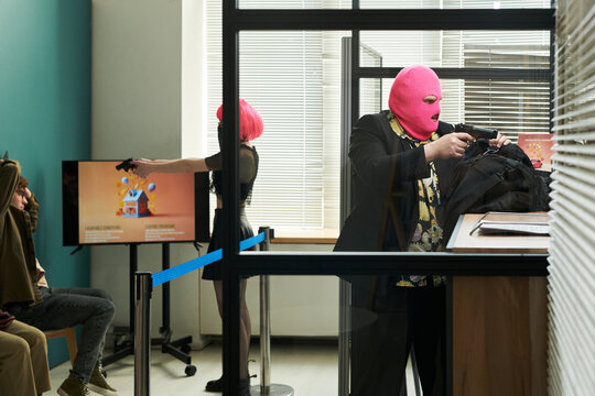 Two young adults wearing pink balaclavas robbing bank, one aiming handgun at seated people while other opening bag at counter