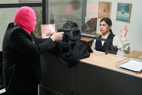 Caucasian man wearing pink ski mask pointing handgun at young adult Hispanic woman behind counter raising hand during bank robbery, office posters and documents visible in background