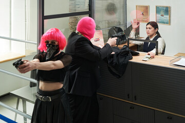 Caucasian young adult woman standing behind counter raising hands while two masked Caucasian young adult individuals pointing handguns during bank robbery scene