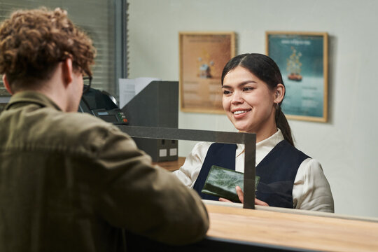 Young adult Hispanic woman smiling while assisting young adult Caucasian man at service counter, holding passport and engaging in customer service interaction through glass partition