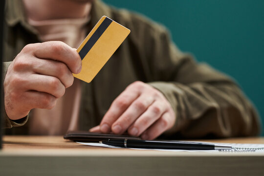 Unrecognizable man holding credit card in right hand while standing at desk with wallet and pen, preparing for online payment or financial transaction, focus on hands